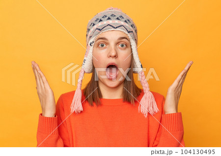 Portrait of astonished young adult Caucasian woman wearing funny earflap hat and orange casual jumper standing isolated over yellow background, raised her arms, being shocked. 104130495