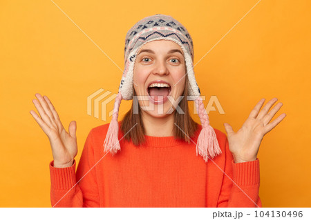 Indoor shot of excited positive happy woman wearing orange jumper and knitted earflap hat standing isolated over yellow background, raised her arms, screaming with happiness. 104130496