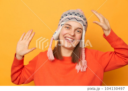 Indoor shot of extremely happy smiling woman wearing funny earflap hat and orange casual jumper standing isolated over yellow background, raised her arms, dancing, celebrating. 104130497