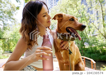 Young charming smiling girl is resting while walking in the park with two golden dogs. The girl hugs her pets. Love and affection between owner and pet. Adopting a pet from a shelter. Young charming smiling girl is resting while walking in the park with two golden dogs. The girl hugs her pets. Love and affection between owner and pet. Adopting a pet from a shelter. 104130574