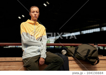 Young fitness girl getting ready for a workout at the stadium. Attractive slim blonde in a yellow-gray sports jacket, khaki cargo pants with a gym bag sits on the edge of the treadmill. 104130884