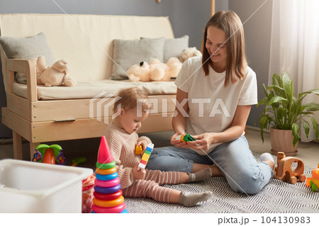 Indoor shot of mother and baby daughter playing with colorful constructor blocks while sitting on the floor in living room at home, family spending time together. Indoor shot of mother and baby daughter playing with colorful constructor blocks while sitting on the floor in living room at home, family spending time together. 104130983