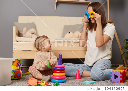 Indoor shot of funny mother wearing white shirt and jeans playing with her little daughter at home while sitting on floor, mommy covering her eyes with rings of pyramid. 104130995