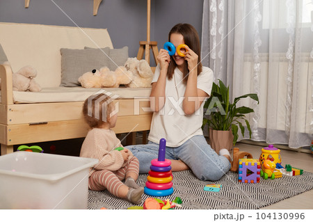 Activities with children at home, early childhood development. Young adult woman sitting on floor in living room, playing with her baby child with pyramid, covering her eyes with rings, having fun. Activities with children at home, early childhood development. Young adult woman sitting on floor in living room, playing with her baby child with pyramid, covering her eyes with rings, having fun. 104130996