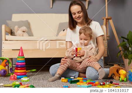Loving Caucasian young mother and cute toddler girl playing with colorful blocks, sitting on warm floor at home, happy mum and adorable little daughter constructing tower, childcare. 104131017