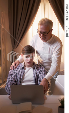 Senior technology. Education help. Computer instructions. Serious young son guy sitting at table and mature dad man standing studying laptop at home in afternoon. Senior technology. Education help. Computer instructions. Serious young son guy sitting at table and mature dad man standing studying laptop at home in afternoon. 104132468