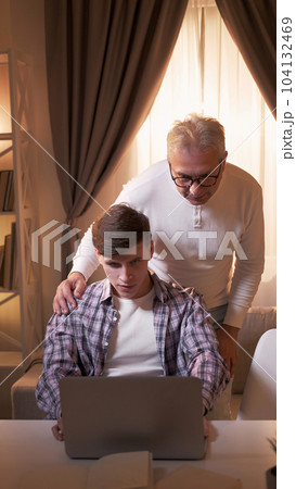 Family technology. Elderly computer. Helping parents. Focused young son guy sitting at table with laptop and grey hair father man standing at home in afternoon. Family technology. Elderly computer. Helping parents. Focused young son guy sitting at table with laptop and grey hair father man standing at home in afternoon. 104132469