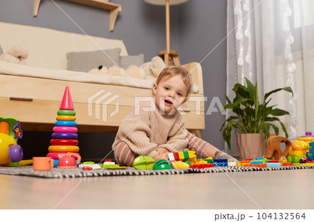 Portrait of cute smiling female kid wearing beige jumper sitting on floor surrounded with colorful toys, looking at camera, playing at home, smiling happily. Portrait of cute smiling female kid wearing beige jumper sitting on floor surrounded with colorful toys, looking at camera, playing at home, smiling happily. 104132564