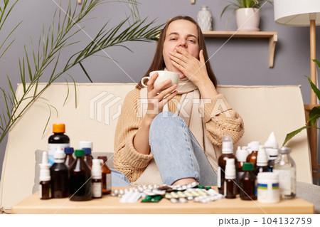 Portrait of sick sleepy woman wearing sweater and wrapped in scarf sitting on sofa at home, drinking hot tea from white cup, yawning, covering mouth with palm. 104132759
