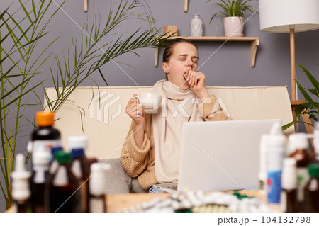 Sleepy exhausted young adult woman sitting on cough in home interior among pills, wearing warm sweater and scarf, yawning and covering her mouth, holding cup with tea, looking at pc computer screen. Sleepy exhausted young adult woman sitting on cough in home interior among pills, wearing warm sweater and scarf, yawning and covering her mouth, holding cup with tea, looking at pc computer screen. 104132798