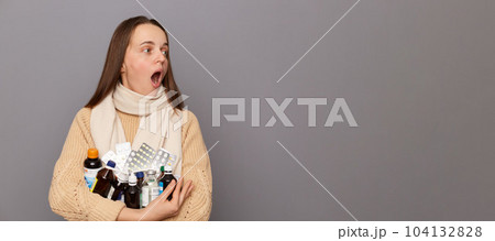 Portrait of surprised shocked caucasian woman wearing jumper and scarf holding pills, looking away at advertisement area, screaming from astonishment, standing isolated over grey background. Portrait of surprised shocked caucasian woman wearing jumper and scarf holding pills, looking away at advertisement area, screaming from astonishment, standing isolated over grey background. 104132828