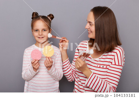 Portrait of cute positive woman and little girl with hair buns wearing casual clothes standing isolated over gray background, kid holding sponges, mommy applying powder with brush. Portrait of cute positive woman and little girl with hair buns wearing casual clothes standing isolated over gray background, kid holding sponges, mommy applying powder with brush. 104133181