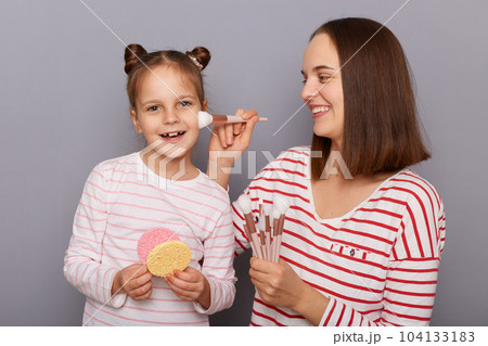 Horizontal shot of smiling young adult brown hair woman with her daughter posing isolated over gray background, mother applying powder with brush to her kid's cheek. 104133183