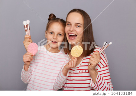 Horizontal shot of excited extremely happy woman with her daughter, holding sponges and cosmetic brushes, looking at camera with joyful expression posing isolated over gray background. 104133184