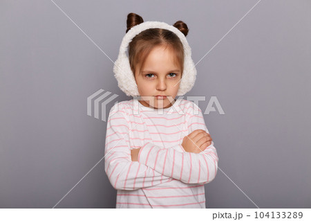 Image of little girl wearing fur earmuffs and striped shirt standing isolated over gray background, being offended, keeps arms folded, looking at cameras with pout lips. Image of little girl wearing fur earmuffs and striped shirt standing isolated over gray background, being offended, keeps arms folded, looking at cameras with pout lips. 104133289