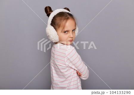 Portrait of sad upset little girl wearing striped shirt and fur headphones standing isolated over gray background, standing backwards to camera, turning with pout lips, being offended. Portrait of sad upset little girl wearing striped shirt and fur headphones standing isolated over gray background, standing backwards to camera, turning with pout lips, being offended. 104133290