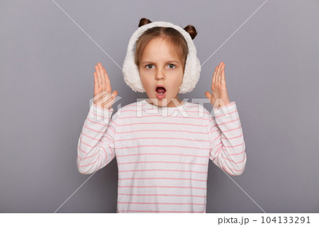 Portrait of shocked astonished little girl wearing striped shirt and fur headphones standing isolated over gray background, raised her arms, sees something shocked, keeps mouth opened. Portrait of shocked astonished little girl wearing striped shirt and fur headphones standing isolated over gray background, raised her arms, sees something shocked, keeps mouth opened. 104133291