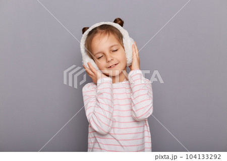 Portrait of relaxed cute charming little girl wearing striped shirt and fur headphones, keeps hands on her ears, posing with closed eyes, enjoying, standing isolated over gray background. Portrait of relaxed cute charming little girl wearing striped shirt and fur headphones, keeps hands on her ears, posing with closed eyes, enjoying, standing isolated over gray background. 104133292