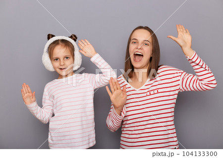 Extremely happy joyful little girl wearing striped shirt and fur headphones standing with her mother isolated over gray background, dancing together, being in festive mood. 104133300