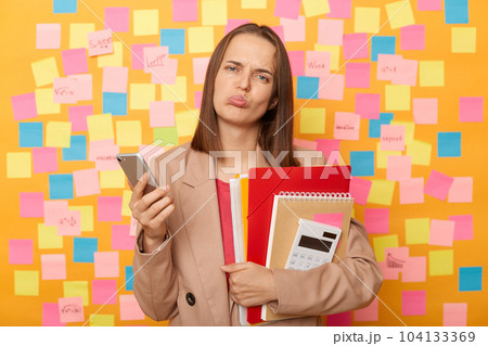Portrait of sad upset disappointed woman wearing beige jacket standing against yellow wall with colorful memo cards, holding paper folders ans calculator, using cell phone, reading bad news. Portrait of sad upset disappointed woman wearing beige jacket standing against yellow wall with colorful memo cards, holding paper folders ans calculator, using cell phone, reading bad news. 104133369