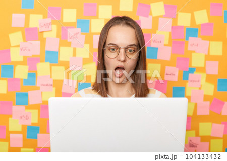 Indoor shot of shocked astonished woman in glasses with brown hair working on laptop against yellow wall with memo cards, looking monitor with open mouth, works online. Indoor shot of shocked astonished woman in glasses with brown hair working on laptop against yellow wall with memo cards, looking monitor with open mouth, works online. 104133432
