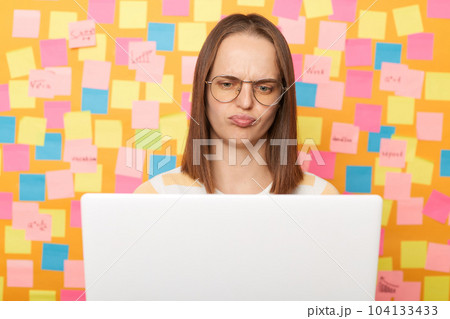 Photo of sad stresses tired brown haired woman wearing T-shirt posing against colorful sticky notes on yellow wall, working on laptop, looking at screen with sad expression. Photo of sad stresses tired brown haired woman wearing T-shirt posing against colorful sticky notes on yellow wall, working on laptop, looking at screen with sad expression. 104133433