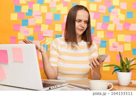 Portrait of confused dark haired woman wearing striped t-shirt sitting at table, posing against multicolored memo cards on yellow wall, looks puzzled, holding mobile phone. 104133511