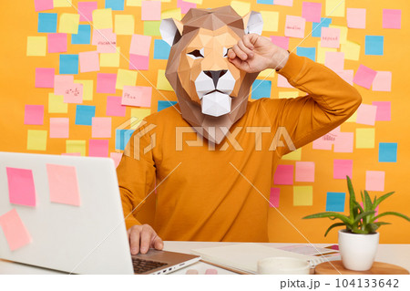 Image of exhausted anonymous man in lion mask wearing orange sweater sitting at workplace with laptop, feels eyes hurt, rubbing her eye, posing against yellow wall with stickers. Image of exhausted anonymous man in lion mask wearing orange sweater sitting at workplace with laptop, feels eyes hurt, rubbing her eye, posing against yellow wall with stickers. 104133642