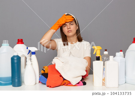 Unhappy young Caucasian woman keeps hand on forehead, having headache, wearing rubber protective gloves and headband, surrounded with bottles of detergents, isolated over gray background Unhappy young Caucasian woman keeps hand on forehead, having headache, wearing rubber protective gloves and headband, surrounded with bottles of detergents, isolated over gray background 104133735
