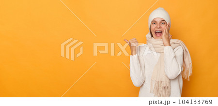 Excited woman wearing white sweater, cap and scarf pointing index fingers on copy space area, looking with excitement and amazement, standing isolated over yellow background. Excited woman wearing white sweater, cap and scarf pointing index fingers on copy space area, looking with excitement and amazement, standing isolated over yellow background. 104133769