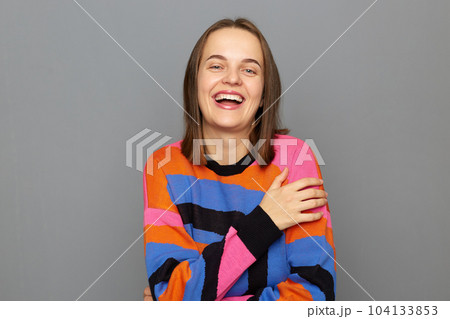 Indoor shot of laughing Caucasian woman wearing colorful sweater posing isolated over gray background, laughs out loud, looking at camera, hearing funny joke. 104133853