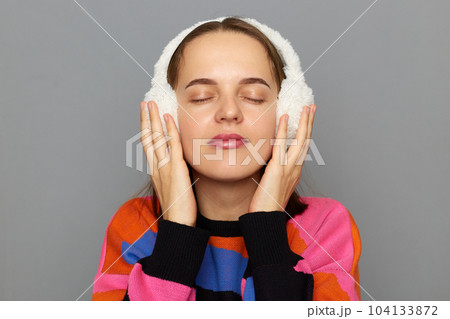 Indoor shot of calm beautiful adorable woman wearing colorful sweater posing isolated over gray background, standing with closed eyes, looks cute, enjoying moment. 104133872
