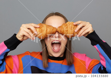 Portrait of funny joyful Caucasian woman wearing sweater posing isolated over gray background, covering her eyes with croissants, keeps mouth widely open, having fun. Portrait of funny joyful Caucasian woman wearing sweater posing isolated over gray background, covering her eyes with croissants, keeps mouth widely open, having fun. 104133880