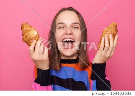 Photo of excited laughing woman with brown hair wearing sweater, holding two croissants in hands, screaming, feels hungry, posing isolated over pink background. Photo of excited laughing woman with brown hair wearing sweater, holding two croissants in hands, screaming, feels hungry, posing isolated over pink background. 104133932