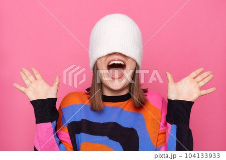 Indoor shot of excited amazed woman wearing jumper pulling white cap on her face and raised arms, screaming happily, being in good mood, expressing positive emotions. 104133933