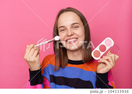 Portrait of smiling delighted woman with dark straight hair wearing jumper standing isolated over pink background, holding palette of eyeshadow in hands, applying powder with brush on her cheek. Portrait of smiling delighted woman with dark straight hair wearing jumper standing isolated over pink background, holding palette of eyeshadow in hands, applying powder with brush on her cheek. 104133938