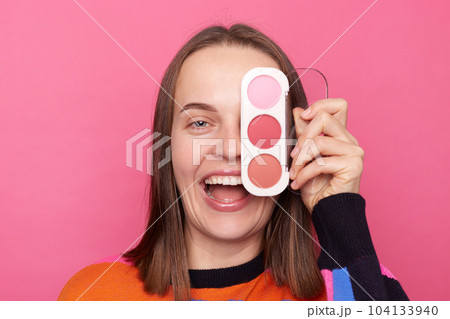 Portrait of young Caucasian woman covering her eye with colorful eyeshadow palette, wearing sweater, doing makeup for dating, posing isolated over pink background. Portrait of young Caucasian woman covering her eye with colorful eyeshadow palette, wearing sweater, doing makeup for dating, posing isolated over pink background. 104133940