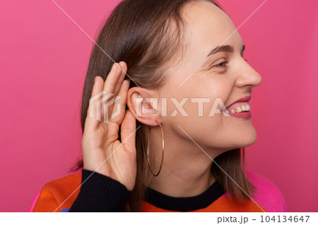 Photo of smiling satisfied woman with brown hair posing isolated over pink background, covering hand near ear, listening to somebody's private conversation, spying, showing earrings. Photo of smiling satisfied woman with brown hair posing isolated over pink background, covering hand near ear, listening to somebody's private conversation, spying, showing earrings. 104134647