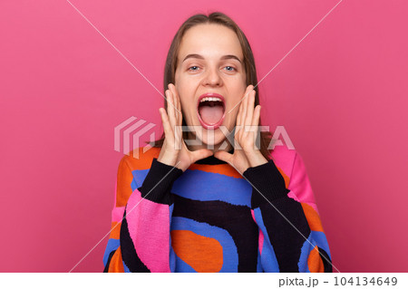 Indoor shot of excited woman wearing sweater posing against pink wall, screaming loud with hands near mouth, making announcement, saying important information. Indoor shot of excited woman wearing sweater posing against pink wall, screaming loud with hands near mouth, making announcement, saying important information. 104134649
