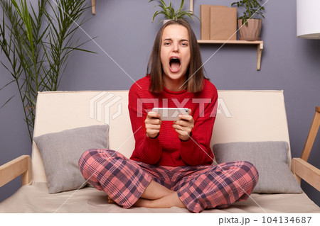 Photo of addicted woman gamer wearing red sweater and checkered pants sitting on sofa in home interior, holding cell phone, lose the level of favorite game, screaming with despair. 104134687