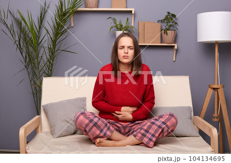 Indoor shot of sick ill brown-haired female wearing red sweater and checkered pants, having unhealthy look, holding her belly, suffering strong stomachache, sitting on cough in room at home. Indoor shot of sick ill brown-haired female wearing red sweater and checkered pants, having unhealthy look, holding her belly, suffering strong stomachache, sitting on cough in room at home. 104134695