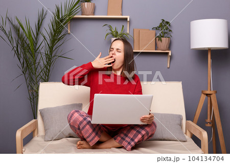 Indoor shot of tired brown-haired female wearing red sweater and checkered pants sitting on cough in room at home, working online on portable computer, feels sleepy, yawning, covering mouth with palm. Indoor shot of tired brown-haired female wearing red sweater and checkered pants sitting on cough in room at home, working online on portable computer, feels sleepy, yawning, covering mouth with palm. 104134704