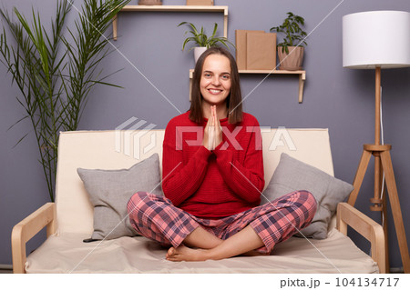 Photo of relaxed friendly pleased Caucasian female dressed in home clothes sitting in lotus pose on cough in her flat, keeps palms in prayer gesture, meditating, smiling to camera, doing yoga. 104134717