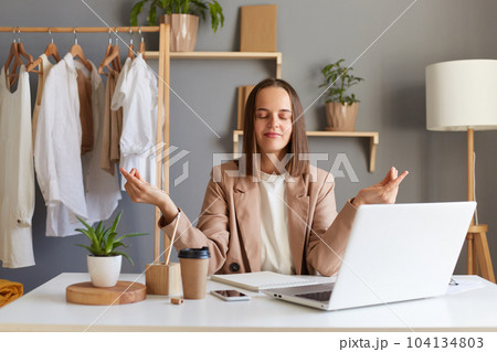 Portrait of relaxed woman wearing beige jacket sitting in front of laptop with clothing on hangers on background, practicing yoga trying to relax and calm down during hard working day. Portrait of relaxed woman wearing beige jacket sitting in front of laptop with clothing on hangers on background, practicing yoga trying to relax and calm down during hard working day. 104134803