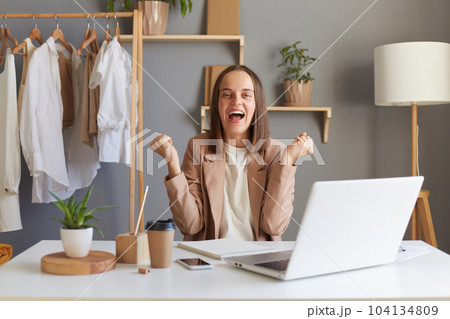 Photo of extremely happy Caucasian brown-haired woman designer wearing beige jacket, posing with clenched fists, celebrating success, sitting at her workplace in showroom in front of laptop. 104134809