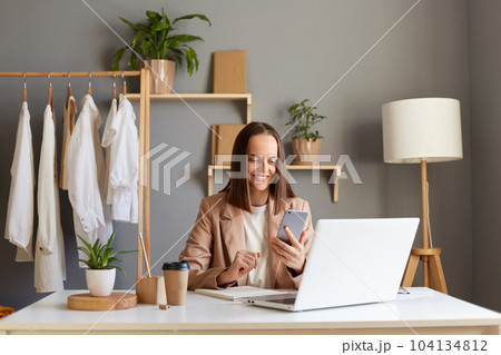 Indoor shot of smiling brown haired woman stylist wearing beige jacket posing in office, posing with clothes on hanger on background, using cell phone, smiling. 104134812