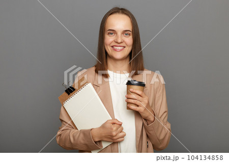 Indoor shot of smiling satisfied brown-haired woman wearing jacket holding clipboard with documents and disposable cup with coffee isolated over gray background. 104134858