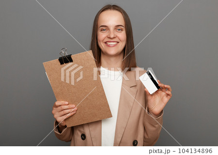 Portrait of positive cheerful woman with brown hair wearing beige jacket, standing with documents and showing credit card, looking smiling at camera, posing isolated over gray background. Portrait of positive cheerful woman with brown hair wearing beige jacket, standing with documents and showing credit card, looking smiling at camera, posing isolated over gray background. 104134896