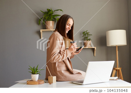 Side view portrait of Caucasian attractive woman with brown hair wearing beige jacket working in office, looking at laptop display, holding smart phone and takeaway coffee. Side view portrait of Caucasian attractive woman with brown hair wearing beige jacket working in office, looking at laptop display, holding smart phone and takeaway coffee. 104135048