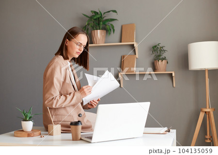 Portrait of young adult attractive businesswoman wearing official style suit holding papers documents and working on laptop, sitting on table in office. Portrait of young adult attractive businesswoman wearing official style suit holding papers documents and working on laptop, sitting on table in office. 104135059